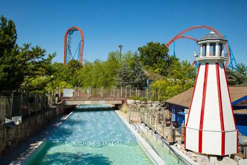 A scenic view of a water attraction with a lighthouse, surrounded by trees and roller coasters in the background.