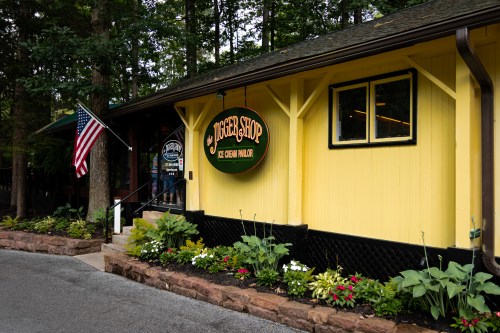 A yellow ice cream shop with a sign reading "Jigger Shop," surrounded by greenery and an American flag.