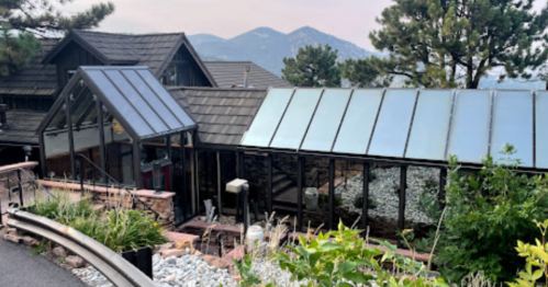 A modern house with large glass windows, surrounded by greenery and mountains in the background.