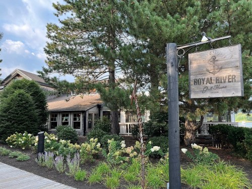 A rustic building with a sign reading "Royal River Grill House," surrounded by greenery and flowers.