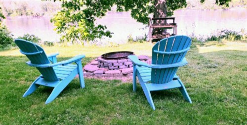 Two blue Adirondack chairs facing a stone fire pit, surrounded by green grass and trees near a body of water.