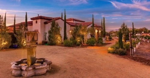 A scenic view of a rustic property with a fountain, surrounded by trees and illuminated by warm evening light.