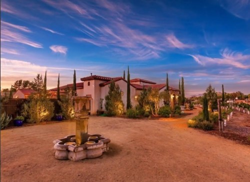 A serene landscape featuring a rustic building, fountain, and cypress trees under a colorful sunset sky.