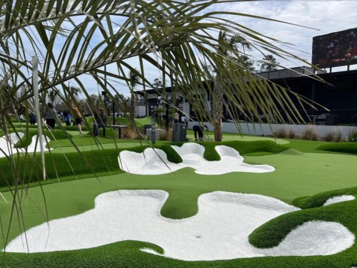 A vibrant mini-golf course with green turf, white sand traps, and palm trees under a sunny sky.