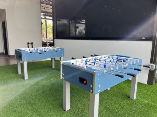 Two blue foosball tables on a green turf surface, set against a modern indoor backdrop.