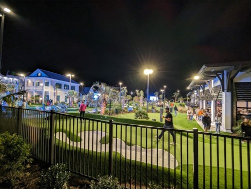 A lively outdoor mini-golf area at night, with people playing and palm trees illuminated by bright lights.