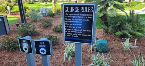 Sign displaying golf course rules with directional signs for black and blue courses, surrounded by greenery.