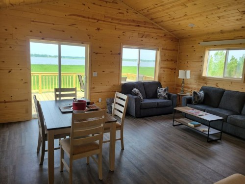 Cozy wooden interior of a lakeside cabin with a dining table, sofas, and large windows overlooking the water.