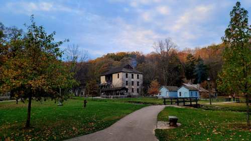 A scenic view of a historic village with stone buildings, surrounded by trees and a pathway under a cloudy sky.
