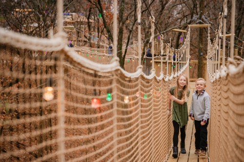 Two children smile on a rope bridge surrounded by trees, with colorful lights decorating the scene.