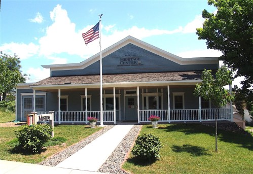 A blue heritage center building with a porch, American flag, and flower planters on a sunny day.