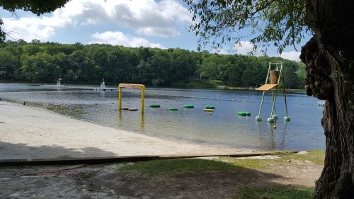 A serene lake scene with a sandy beach, floating obstacles, and a lifeguard chair under a blue sky with clouds.