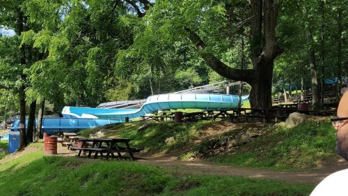 A water slide in a wooded area, surrounded by picnic tables and greenery, with a clear blue sky above.