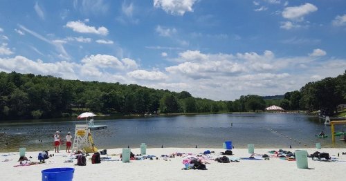 A sandy beach by a lake with people, lifeguard stand, and colorful umbrellas under a blue sky with fluffy clouds.