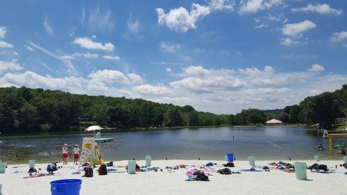 A sunny beach scene with a lake, sandy shore, scattered towels, and trees in the background under a blue sky with clouds.