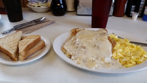 A plate of biscuits and gravy with scrambled eggs, alongside a side of toast on a white table.