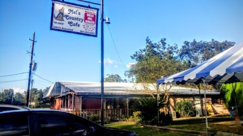 A rustic cafe building with a sign reading "Mel's Country Cafe" and a striped tent nearby, set against a clear blue sky.