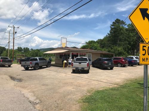 A small roadside eatery with several parked trucks and cars, surrounded by greenery and under a blue sky.
