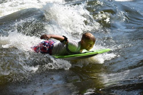 A child rides a green boogie board through a wave, splashing water around in a sunny outdoor setting.