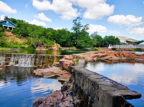 A serene landscape featuring a waterfall, calm water, rocky shore, and lush greenery under a blue sky with fluffy clouds.