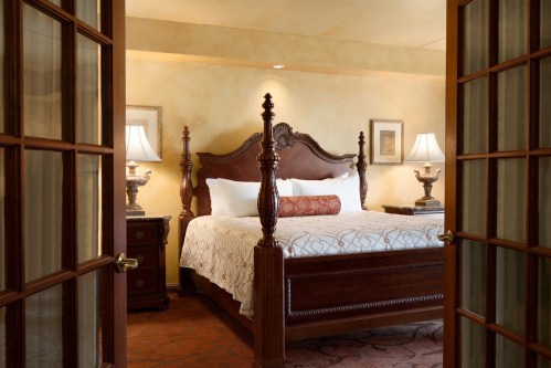 A cozy bedroom with a wooden bed, decorative lamps, and warm-toned walls, viewed through double doors.