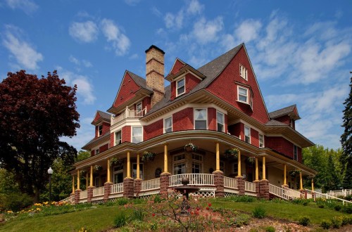 A large, red Victorian-style house with a wraparound porch, surrounded by lush greenery and colorful flowers under a blue sky.