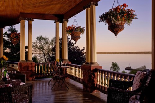 A serene porch with hanging flower baskets, overlooking a calm lake at sunset, featuring cozy seating and a table.