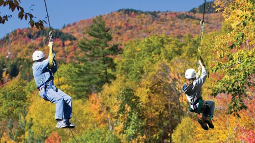 Two people ziplining through a colorful autumn forest with vibrant trees in the background.