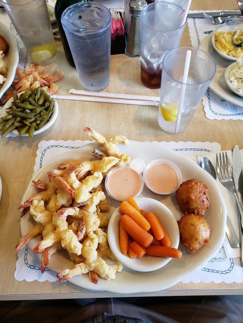 A plate of fried shrimp with dipping sauce, carrots, and hushpuppies, surrounded by drinks and side dishes on a table.