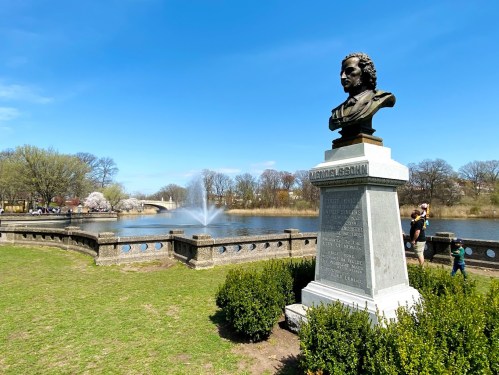 A bust of Mendelssohn stands by a pond with a fountain, surrounded by greenery and trees under a clear blue sky.