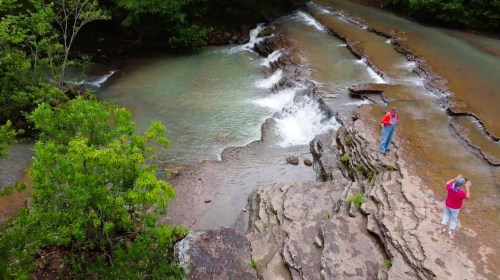 Two people stand by a rocky stream, surrounded by greenery, with water flowing over the rocks in a serene landscape.