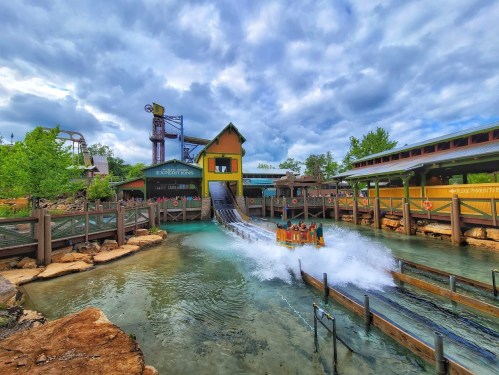 A water ride splashes down at an amusement park, surrounded by greenery and a cloudy sky.