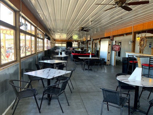 Interior of a restaurant with marble-top tables, black chairs, and a bar area, featuring large windows and ceiling fans.