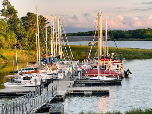 A marina with several sailboats docked at sunset, surrounded by lush greenery and calm waters.