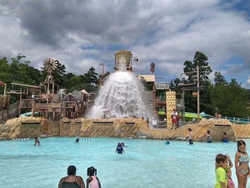 A water park scene with a large water slide and a splash area, featuring children playing in the pool under a cloudy sky.