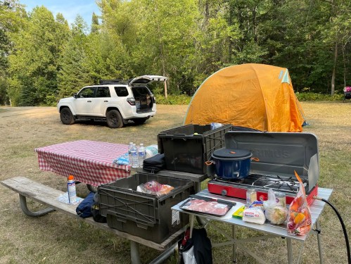 A campsite with a picnic table set up with food, a cooking pot, and a tent in the background next to a parked SUV.