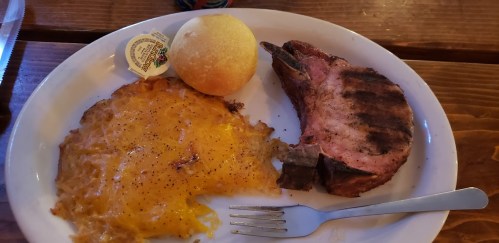 A plate with grilled pork chop, cheesy hash browns, a roll, and a butter packet on a wooden table.