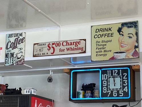 A wall display featuring humorous signs about coffee, whining charges, and a vintage woman enjoying a cup of coffee.