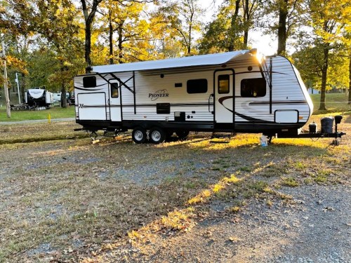 A parked travel trailer in a wooded area, surrounded by autumn foliage and soft sunlight.