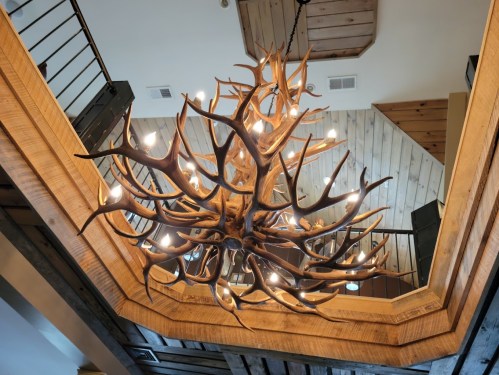 A large antler chandelier hangs from a wooden ceiling in a rustic interior setting.