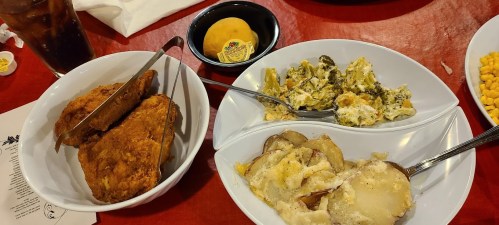 A table setting with fried chicken, broccoli casserole, cheesy potatoes, and a biscuit on a red tablecloth.