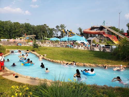 A busy water park scene with people floating in a lazy river, water slides, and lounge chairs under blue umbrellas.