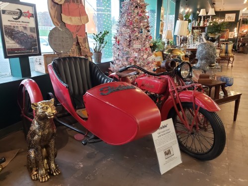 A vintage red motorcycle with a sidecar, displayed in a museum alongside a decorative leopard statue and a Christmas tree.