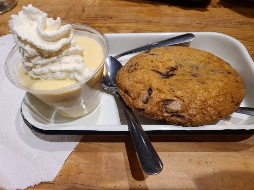 A chocolate chip cookie next to a cup of vanilla pudding topped with whipped cream, served on a tray with spoons.