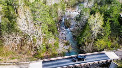 Aerial view of a car driving over a bridge above a winding creek surrounded by trees.