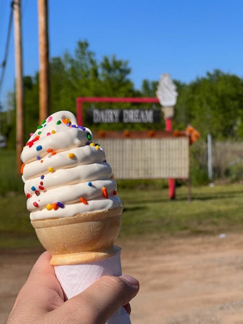 A hand holds a soft-serve ice cream cone with sprinkles, with a "Dairy Dream" sign in the background.