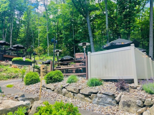 A scenic outdoor dining area surrounded by trees, featuring tables with umbrellas and a stone pathway.