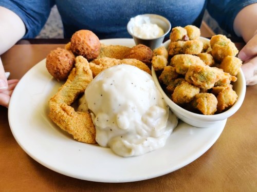 A plate of fried catfish, creamy mashed potatoes, fried okra, and hushpuppies, served with a small cup of sauce.