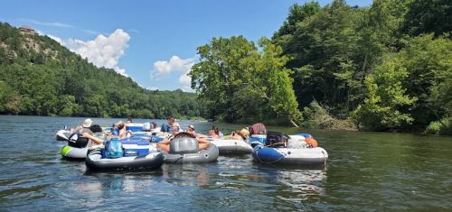 Group of people relaxing on inflatable rafts in a river, surrounded by lush greenery and blue skies.