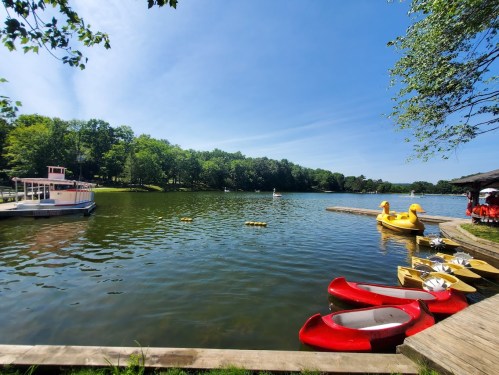 A serene lake scene with colorful kayaks and a paddle boat, surrounded by lush greenery under a clear blue sky.